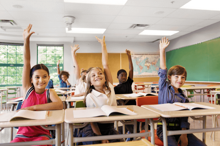 A group of elementary school children sitting at desks with open books, eagerly raising their hands to answer a question.