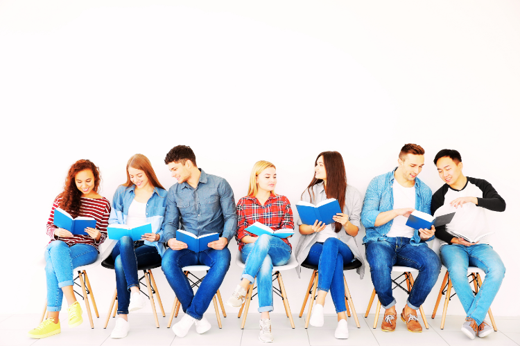 Group of young adults sitting in a row of chairs, reading blue-covered books and discussing together.