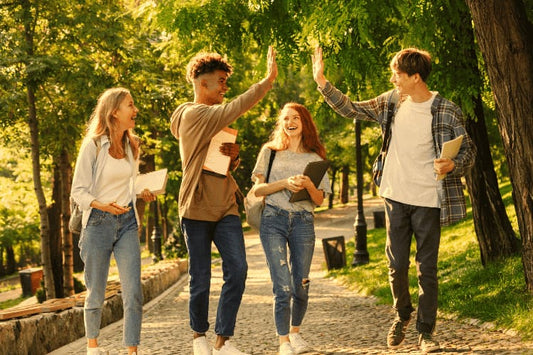 Four happy students walking together in a park, holding notebooks, with two giving each other a high five.