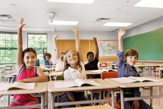 A group of elementary school children sitting at desks with open books, eagerly raising their hands to answer a question.