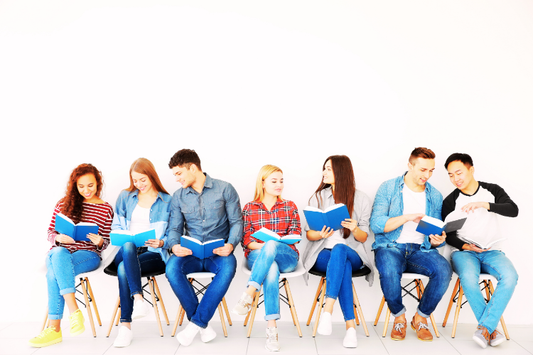Group of young adults sitting in a row of chairs, reading blue-covered books and discussing together.